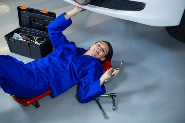 Female mechanic lying on a creeper and smiling while repairing a car in a garage. She is holding a wrench and surrounded by various tools. Ideal for use in automotive service promotions, mechanic training materials, and advertisements for car repair services.