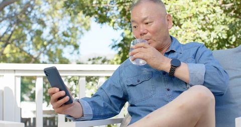 Mature man relaxing on sunlit patio checking smartphone while sipping water
