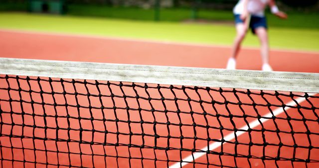 This vibrant image shows a tennis player in action on a clay court, focusing on the tennis net in the foreground. Ideal for websites, blogs, and articles related to sports, tennis tutorials, fitness, and outdoor activities. Great for promotional materials for tennis clubs and sport events.