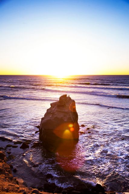 Rock formation standing in the sea during a vivid sunset with lens flare. Waves gently crash against the rock and reflect the golden light of the setting sun, creating a serene and tranquil scene. Ideal for backgrounds, nature themed projects, travel promotions, and relaxation concepts.