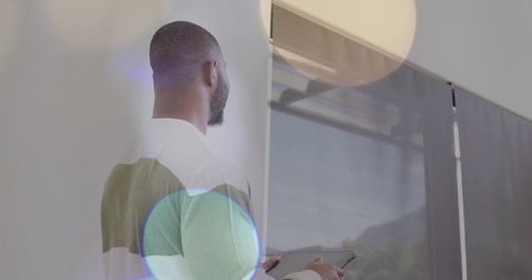 Backview man standing holding tablet by glass window with dark curtains and lens flare