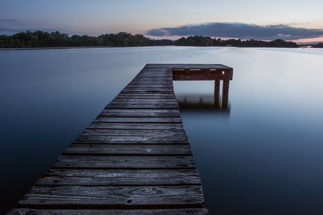 This image showcases an empty, weathered wooden dock extending over still waters at twilight. The serene and tranquil scene captures the calming effect of nature with the soft light of dusk reflecting on the water's surface. Ideal for use in travel brochures, meditation apps, relaxation websites, or as a background image for presentations emphasizing peace and tranquility.