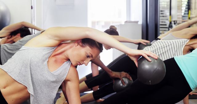 Young women practicing Pilates exercises with medicine balls in a modern fitness studio. They are in a group class focusing on building strength, flexibility, and balance. This stock photo can be used for advertising fitness programs, illustrating workout routines, promoting health and wellness campaigns, or creating content for lifestyle blogs.