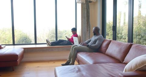 African american father working on laptop while son using smartphone on window seat