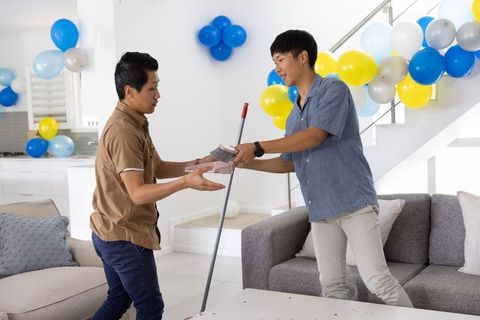 Asian friends cleaning up confetti after home party, passing broom and dustpan by sofa
