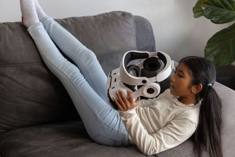 Asian girl reclining on gray sofa inspecting white vr headset in modern living room