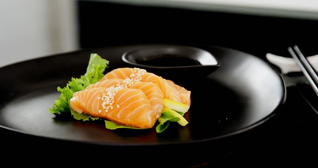 Slices of fresh salmon sashimi are elegantly presented on a black plate, garnished with lettuce and sprinkled with sesame seeds. Sashimi is a popular Japanese delicacy consisting of fresh, raw fish, often enjoyed for its delicate taste and texture.