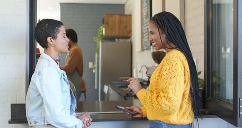 Diverse female friends chatting at cafe counter, holding phone, smiling and connecting