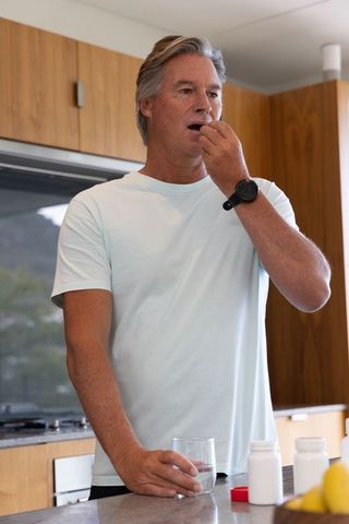Mature man taking pill at kitchen counter holding glass wearing smartwatch pastel t-shirt