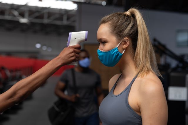 Caucasian woman wearing a blue mask having her temperature checked with a thermal scanner inside the gym. in the background is a man wearing a mask about to leave the gym.