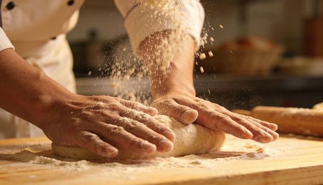 Artisan baker kneading dough as flour dusts over wooden board, closeup hands