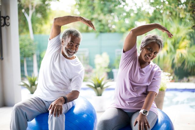 Senior couple sitting on fitness balls in yard, stretching and smiling. Ideal for promoting active lifestyles, healthy aging, and fitness routines for elderly. Suitable for wellness programs, retirement community advertisements, and health-related articles.
