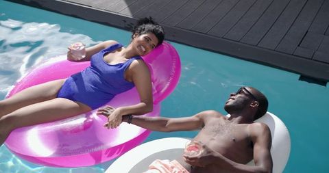 Young couple floating on bright pool inflatables holding hands and sipping cold drinks