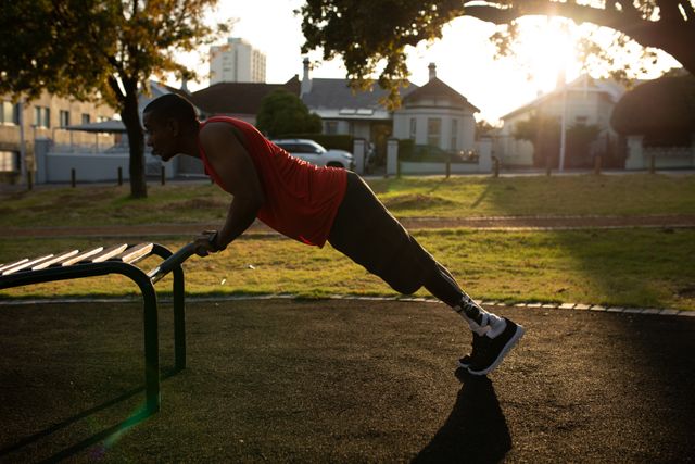 Man with a prosthetic leg doing push ups at an outdoor gym in a park during sunset. This image can be used to promote fitness, healthy lifestyle, and motivation for people with disabilities. It is ideal for articles, advertisements, and social media posts related to adaptive sports, physical rehabilitation, and inclusive fitness programs.