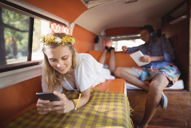 Young woman lying on bed in camper van, using smartphone, smiling. Man in background, sitting, holding digital tablet, wearing headphones. Ideal for travel blogs, vacation advertisements, lifestyle magazines, and technology use in leisure settings.