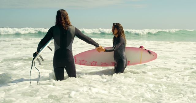 Two young Caucasian women in wetsuits are holding hands and carrying a surfboard at the beach, with copy space. Their shared activity suggests a close bond and a passion for surfing.