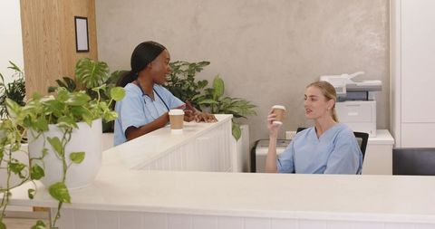 Nurses chatting and sipping coffee at clinic reception surrounded by plants and calm decor