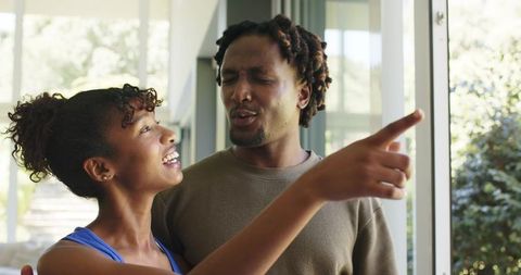African american couple sharing laugh pointing toward window in sunlit contemporary home
