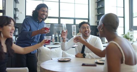 Diverse friends clinking wine glasses and cups around sunlit round table in modern kitchen