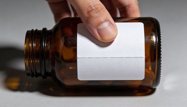 Hand pressing blank label on amber pill bottle closeup for pharmaceutical packaging mockup