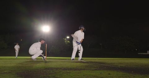 Diverse male cricketers playing on floodlit night pitch batting and wicketkeeping action