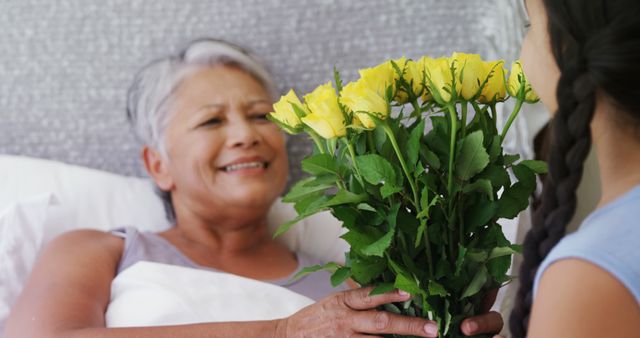 A young girl is presenting a bouquet of yellow roses to a smiling senior woman lying in bed, with copy space. This tender moment captures the care and affection shared between generations, within a family setting.