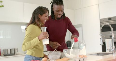 Multicultural couple baking together in bright kitchen pouring milk into mixing bowl