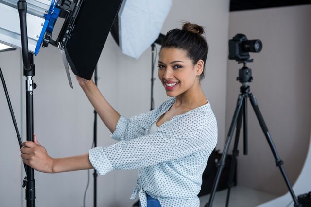 Young female photographer adjusting a spotlight in a professional studio. She is smiling and appears to be enjoying her work. The studio is equipped with various camera equipment and lighting setups. This image can be used for articles or advertisements related to photography, creative professions, or studio setups.