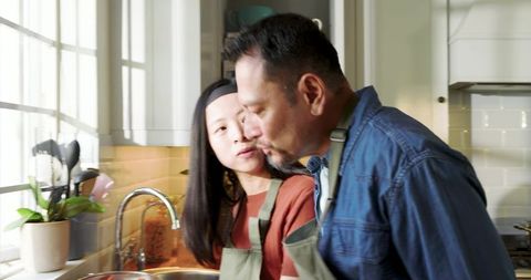 Asian couple cooking together in sunlit kitchen wearing green aprons sharing meal prep
