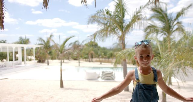 Young girl enjoying her vacation at a tropical resort full of palm trees and bright sunshine. She is smiling joyfully, wearing sunglasses and denim overalls. Ideal for travel brochures, family holiday promotions, summer travel advertisements, and children's vacation planning materials.