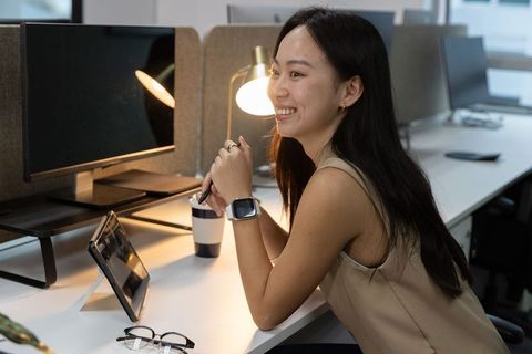 Asian professional woman smiling leaning at desk holding pen using tablet in modern office