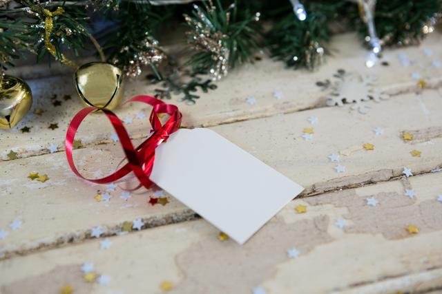 Blank gift tag with red ribbon lying on rustic wooden surface, surrounded by Christmas tree decorations including bells and stars. Ideal for holiday greeting cards, festive invitations, or seasonal marketing materials.