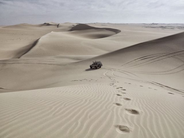 Shows an off-road vehicle traversing vast desert dunes, with footprints leading towards it. Perfect for themes related to adventure, travel, exploration, and uncharted territories. Ideal for use by travel agencies, adventure tour businesses, and nature enthusiasts showcasing rugged landscapes.