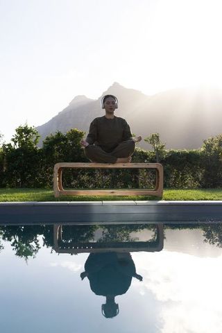 Asian woman meditating with headphones by pool at sunrise with mountain reflection