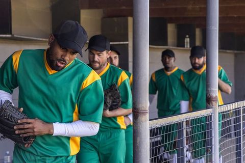 Baseball players preparing in dugout, lead player inspecting glove while teammates lining up