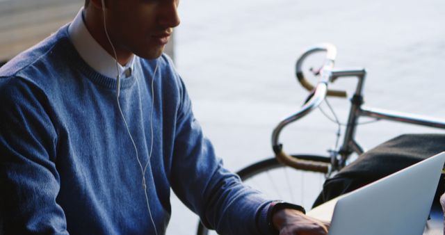 Young man in blue sweater working on laptop while listening to earphones, with a bicycle in the background. Ideal for concepts of remote work, urban lifestyle, modern technology use, productivity, and balancing work and health.