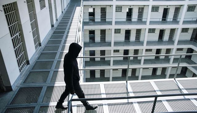 Hooded figure walking grated catwalk inside multi-level prison atrium, metal walkways