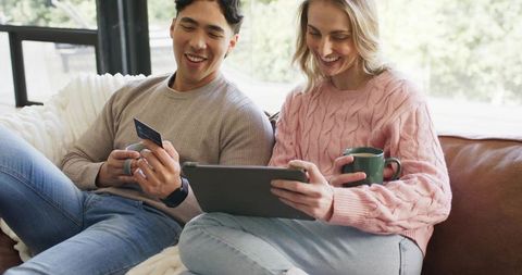 Diverse couple shopping online together on tablet with credit card and coffee, cozy home