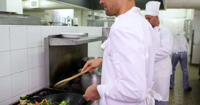 Chefs dressed in white uniforms are diligently preparing food in a professional restaurant kitchen. The main focus is a chef stirring vegetables in a frying pan on the stove, highlighting teamwork and the high standards of the culinary arts. Suitable for articles on culinary education, restaurant operation, teamwork in kitchens, and promotional material for culinary schools and restaurant businesses.