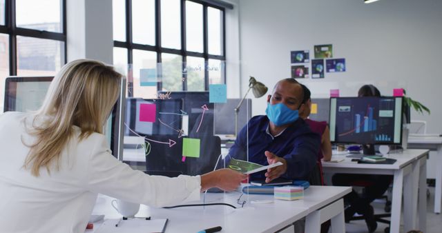 Colleagues in a modern office are collaborating while adhering to pandemic safety measures, such as clear dividers and masks. This setup is ideal for illustrating themes of teamwork, communication, and adaptation within professional environments during pandemic times. Images like this can be used in articles, websites, and presentations discussing pandemic adaptations in the workplace and collaborative efforts.