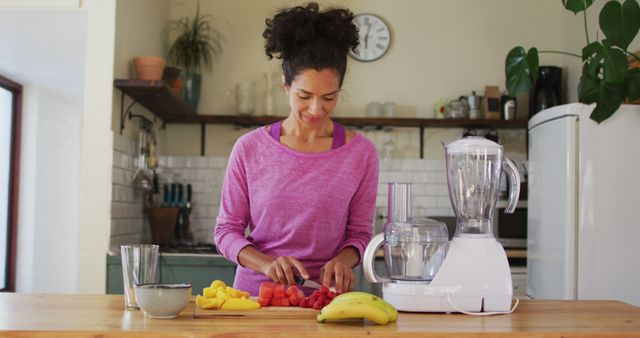 Woman wearing pink shirt cutting fresh fruits including watermelon, pineapple, and bananas on wooden counter. Blender and kitchen tools visible in kitchen. Ideal for content related to healthy living, meal prep, cooking tutorials, kitchen appliances, or lifestyle blogs.