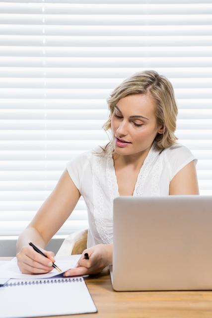 Young woman sitting at home desk writing on paper, with a laptop in front of her. She appears focused and professional, suitable for themes related to remote work, studying, freelancing, education, and productivity.
