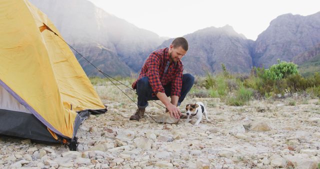 Man enjoying outdoor camping along with a small dog near mountains. Useful for promoting camping, adventure tourism, outdoor activities, and pet-friendly travel. Ideal for advertising on social media, blogs, travel websites, and posters encouraging outdoor experiences and trips.