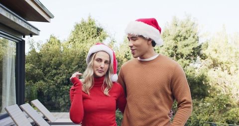 Multicultural couple wearing santa hats sharing warm holiday moment on sunny deck