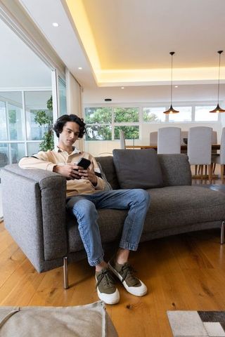 Young man relaxing on gray sofa scrolling smartphone in modern open-plan living room
