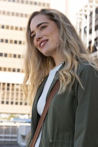 Smiling young woman wearing olive jacket white top carrying brown crossbody bag urban daylight