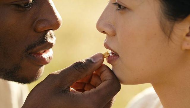 Sharing gentle bite and tender gaze during sunlit meadow picnic, romantic intimacy