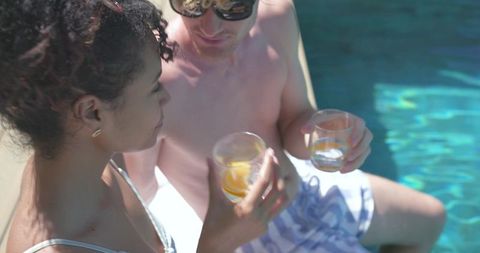 Young couple sipping orange cocktails poolside, relaxing in sunglasses and swimwear