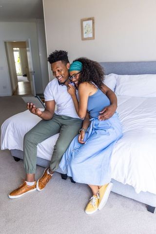 Laughing black couple cuddling on bed browsing tablet, cozy minimalist bedroom romance