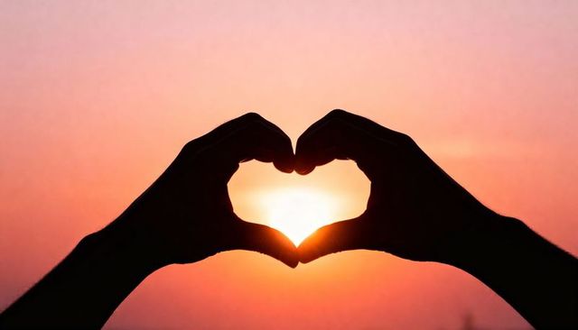Silhouetted hands forming heart framing sun at sunset warm glow romantic sky silhouette